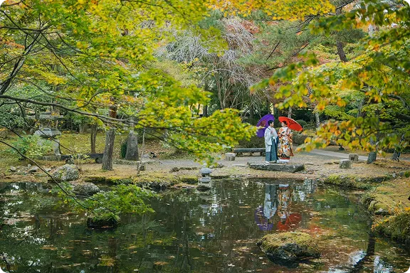 日本庭園で池を前に和装フォトウェディング｜新郎新婦が紫と赤の和傘を手に向き合っている